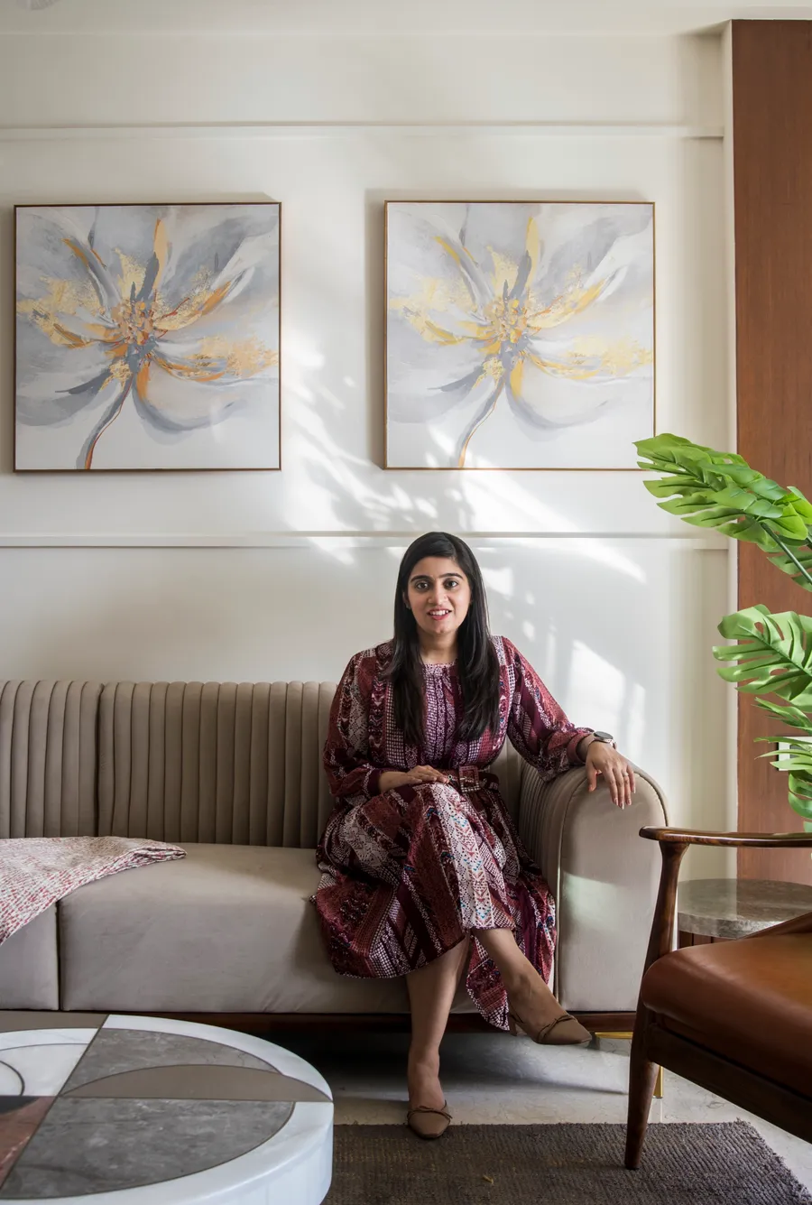Nikhita Chawla seated on a beige channel-tufted sofa beneath two floral paintings