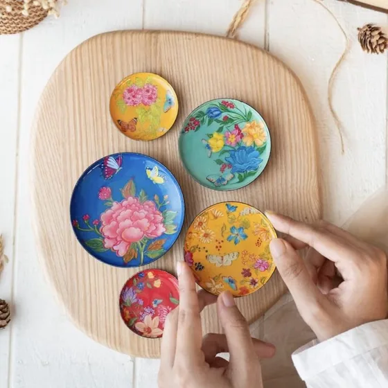 Five small hand-painted floral plate magnets on a wooden board, a hand arranging them