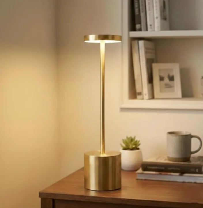 A slim brass cordless table lamp lit on a wooden chest beside a mug and a small plant