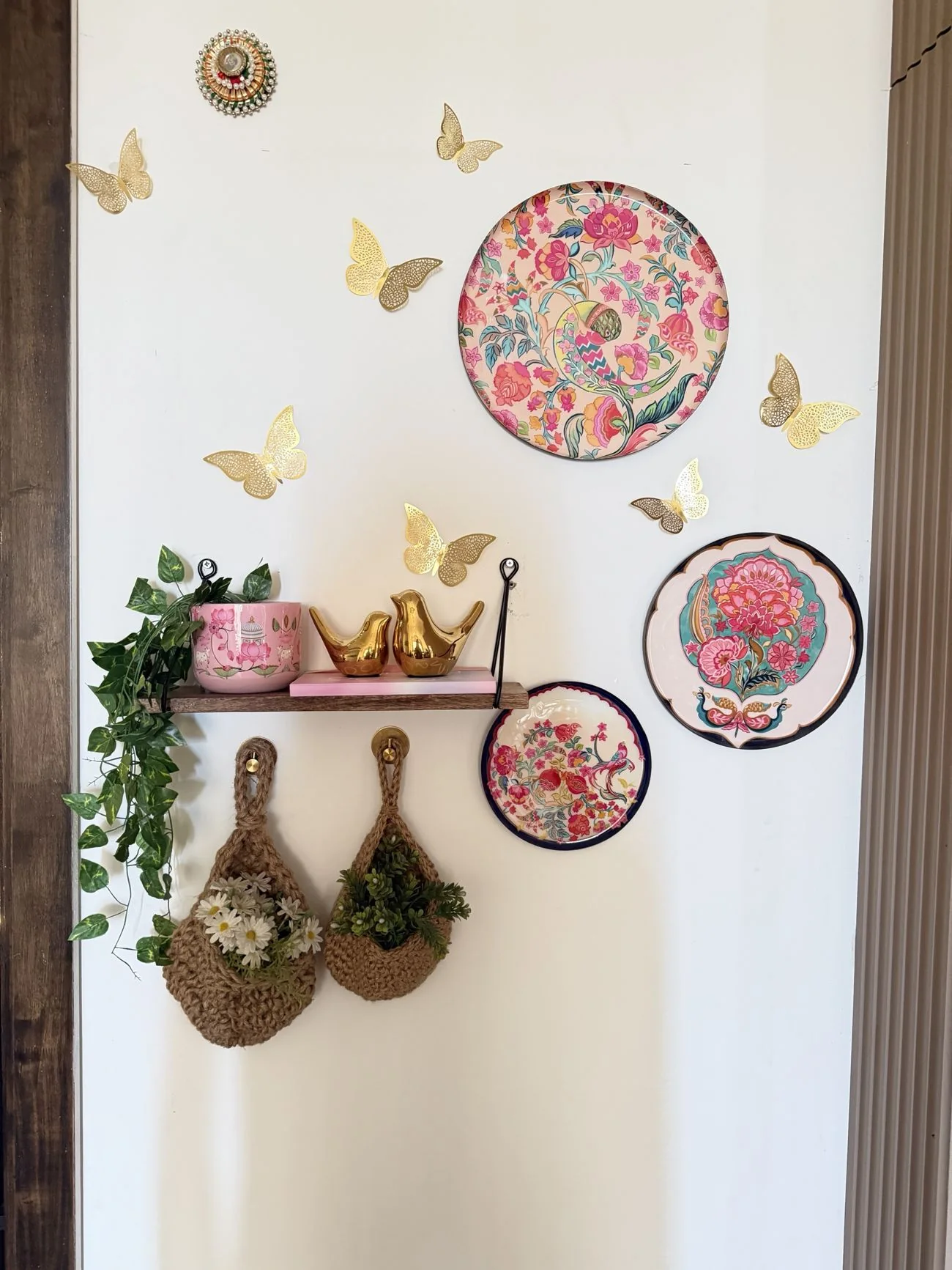 A white wall styled with painted floral plates, gold filigree butterflies, a wooden rope-hung shelf holding a pink planter and gold birds, and jute hanging baskets